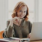 Confident joyful redhead woman works on modern laptop, poses in coworking space, wears sweater