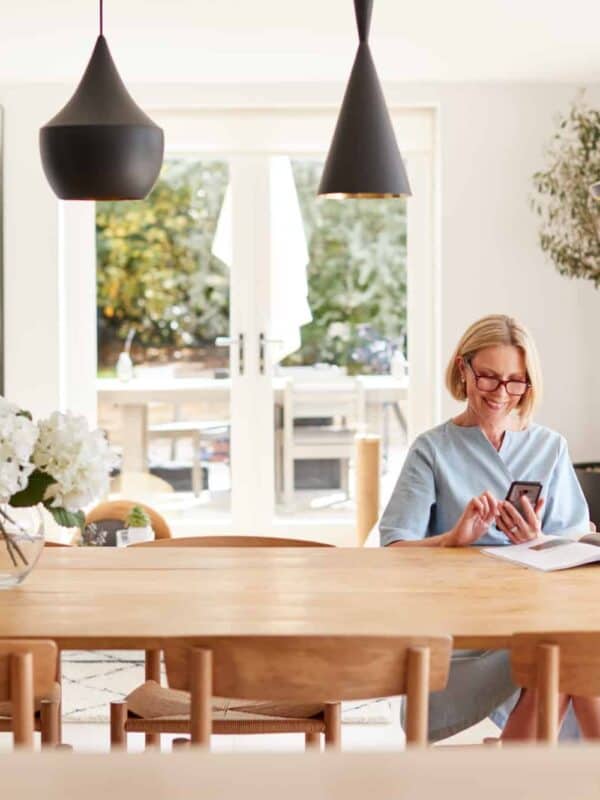 Senior Woman Relaxing With Magazine At Home Looking At Mobile Phone Sitting At Dining Room Table